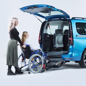 Behindertengerechtes Auto blauer VW Caddy mit geöffnetem Kofferraum und Passivfahrerin im Rollstuhl, einer Frau im Rollstuhl, die über eine Auffahrrampe ins Auto geschoben wird